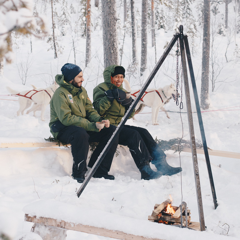 Two people sitting on a bench in the snow close to a fire. Husky sled dogs in the background.