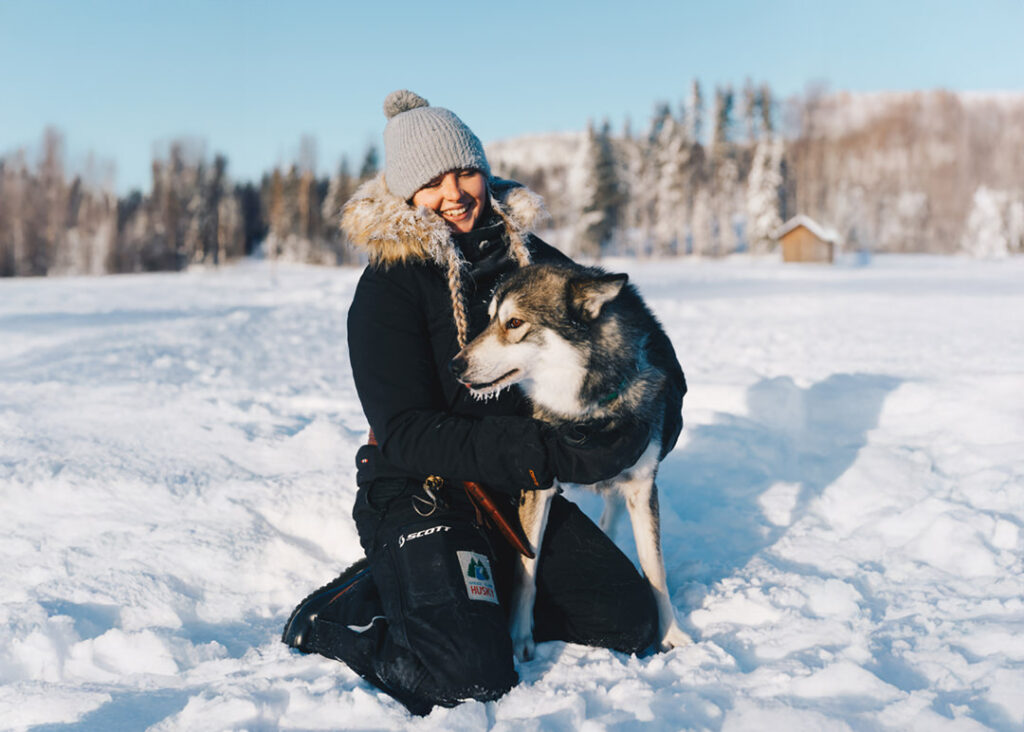 A young woman sitting in the snow with a husky dog.