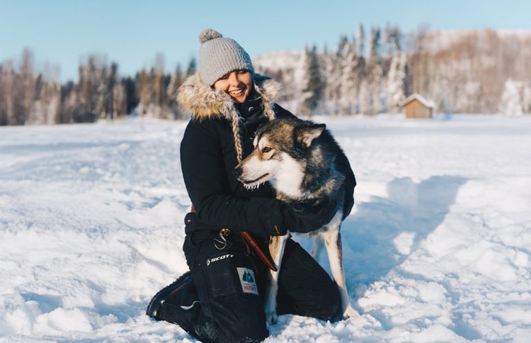 A young woman sitting in the snow with a husky dog.